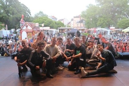 Bild: Gruppe von Jugendlichen sitzt auf dem Boden. Im Hintergrund ist ein Volksfest zu sehen.